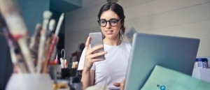 Mujer joven sentada frente a la pantalla de un computador mirando un celular